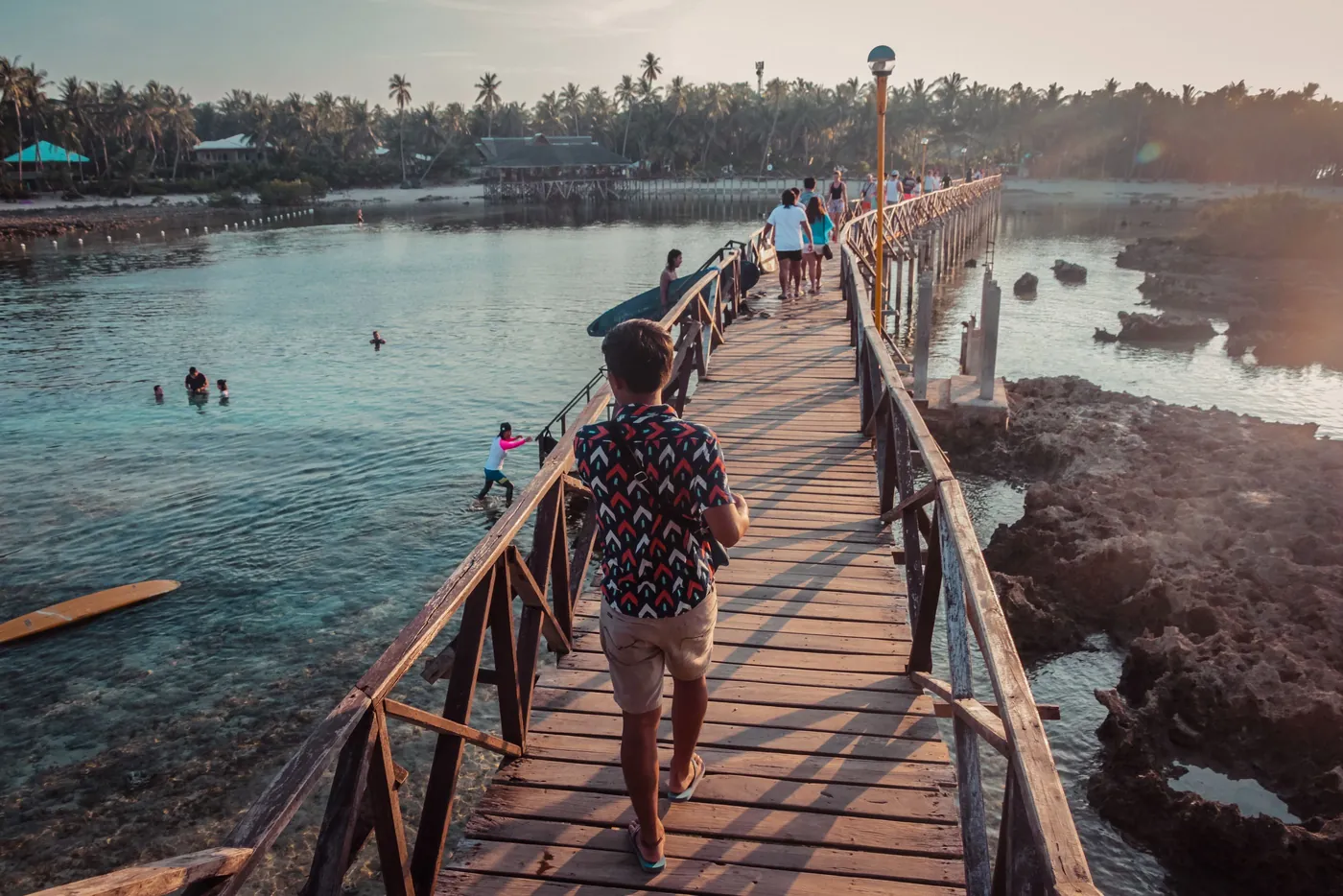 Photo: John Matthew (Pexels License) People walking across a wooden bridge over water at sunset in General Luna, Siargao