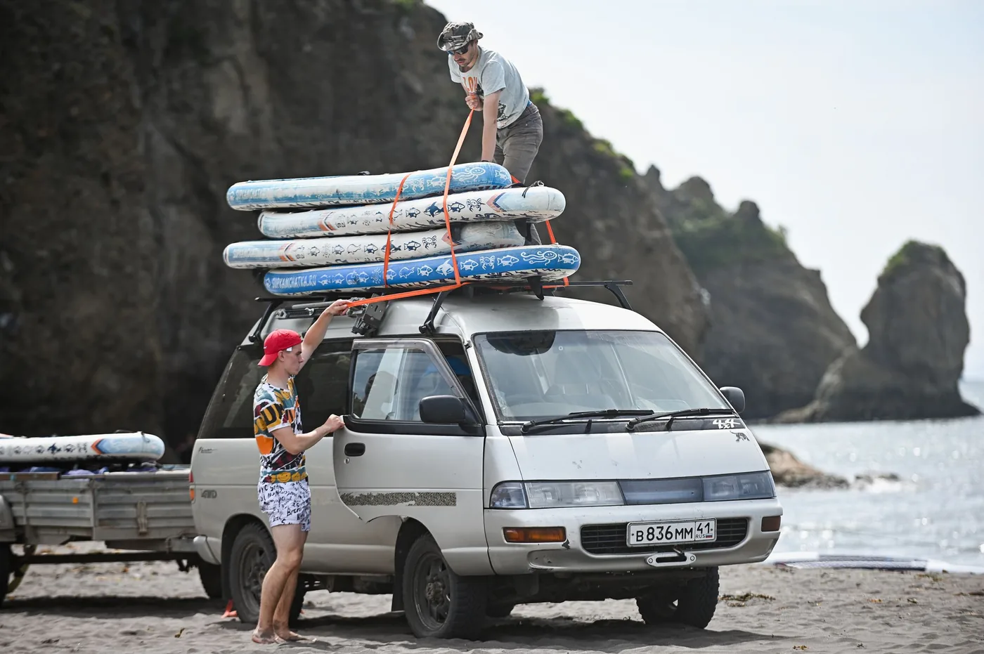 Photo: Dmitrii Eremin (Pexels License) Two people lifting surfboards onto the roof rack of a van parked beside a beach